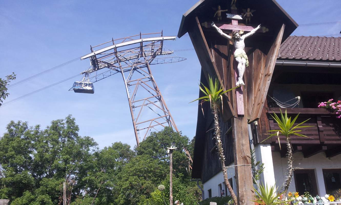 Bildtitel: holzgeschnitzes Kruzifix am Wegesrand mit Blick auf die Pfänderseilbahn